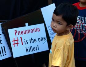 Child stands in front of a sign reading, "Pneumonia #1 is the one killer!" in the Philippines